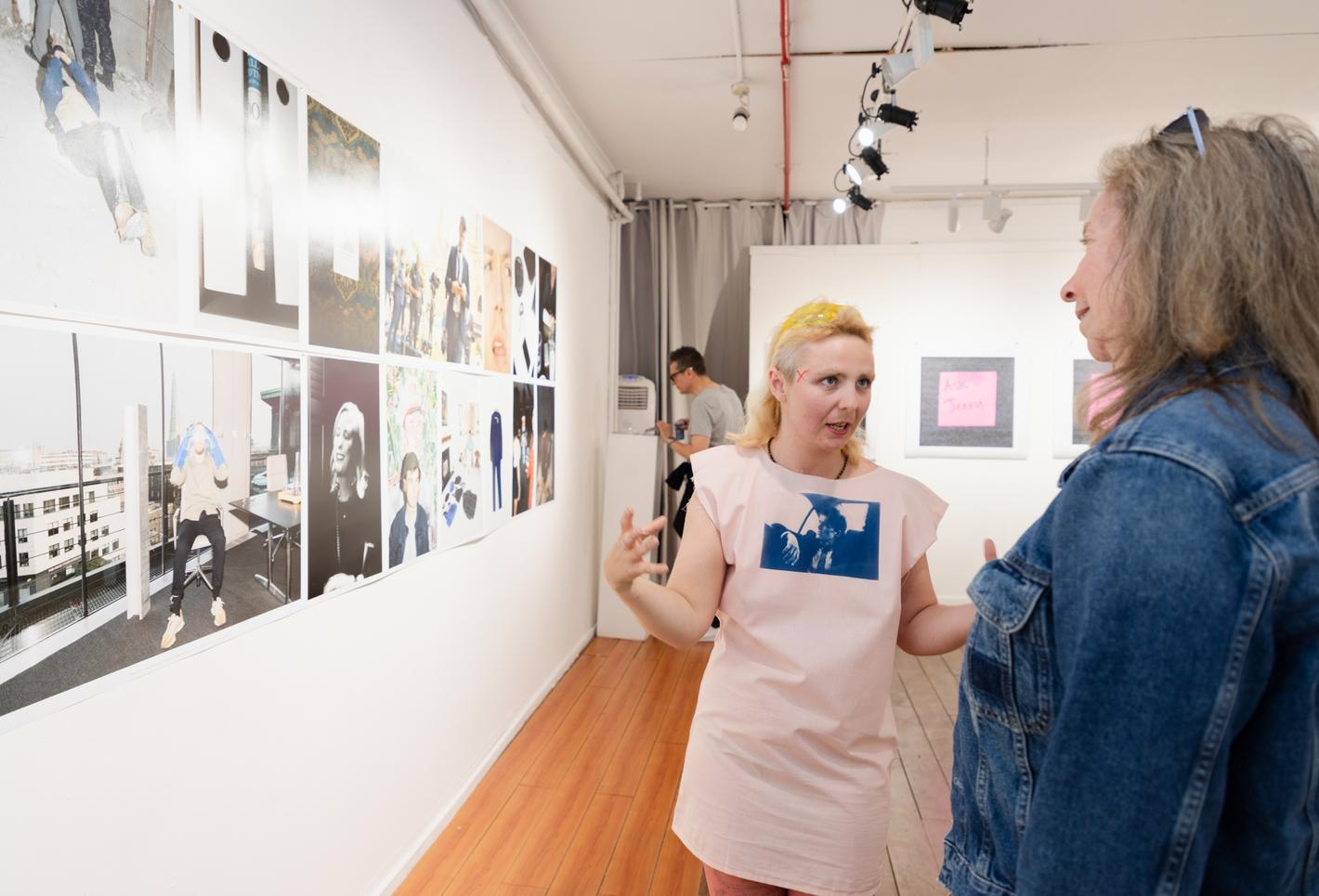A short, young white woman with blond hair in a white shirt dress talks to midle-aged woman with long greyish hair in a denim jacket. They are at a photography exhibition, with work on display on the wall to the left