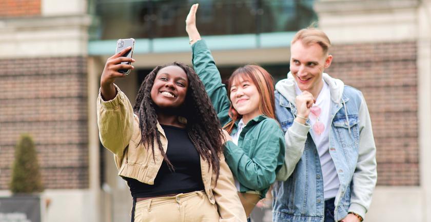 Three young people smiling and posing for a selfie in front of a university building.