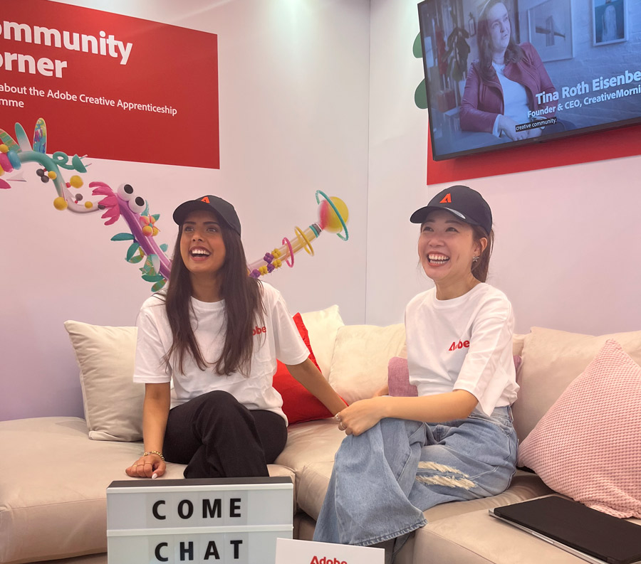 Two young women in white tops and baseball caps sit on a sofa. A sign in front of them reads