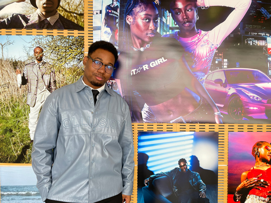A young back man in a grey jacket and black glasses stands in front of large vivid fashion photos displayed on a wall